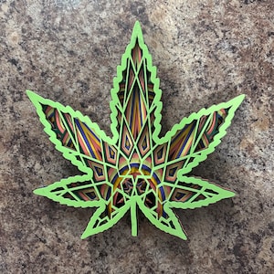 Könnte beinhalten: Ein grünes, geschichtetes, geometrisches Cannabisblatt-Design mit komplizierten Mustern in Rosa-, Gelb-, Blau- und Grüntönen.
