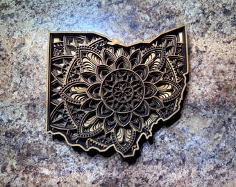 May include: A wooden cutout of the state of Ohio with a detailed mandala design in the center. The mandala is made up of intricate patterns and shapes, and it is a beautiful example of laser-cut art.