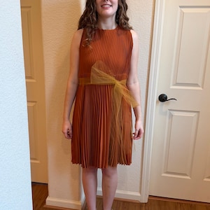 May include: A sleeveless, pleated rust-colored dress with a mustard-yellow bow at the waist. The dress has a knee-length hemline and is paired with nude-colored heels. The dress is suitable for a special occasion.