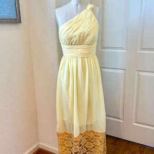 May include: A pale yellow, one-shoulder dress with a pleated bodice and a fitted waist. The skirt flows to the floor and features a golden lace hem. The dress is displayed on a mannequin.