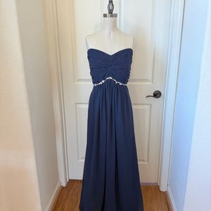 May include: A navy blue strapless formal gown on a mannequin. The bodice is gathered and features a beaded embellishment at the waist. The floor-length skirt flows gracefully. The dress is displayed in front of a white door.