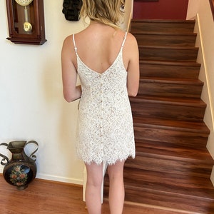 May include: A white lace dress with a v-neckline and spaghetti straps. The dress has a button closure down the back and a scalloped hem.