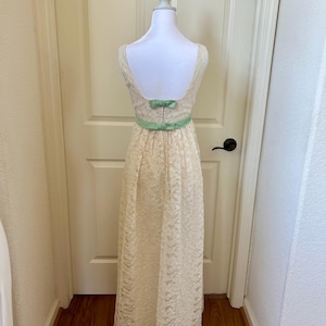 Puede incluir: Vestido de encaje color crema con espalda en forma de cuchara y cinturón de cinta verde claro con lazos. El vestido se exhibe en un maniquí blanco frente a una puerta blanca.
