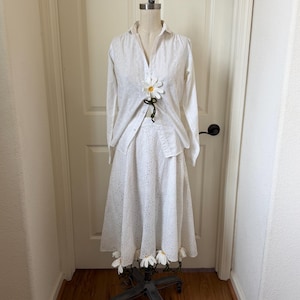 May include: A white eyelet skirt and long-sleeved shirt outfit on a dressmaker's dummy. The shirt is partially unbuttoned, with a large daisy accent. The skirt has daisy appliqués at the hem. The outfit is displayed on a black metal dress form.