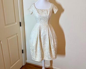 May include: A cream-colored dress with a subtle metallic floral pattern, featuring short sleeves, a square neckline, and a structured, pleated skirt that falls to the knee, displayed on a white mannequin.