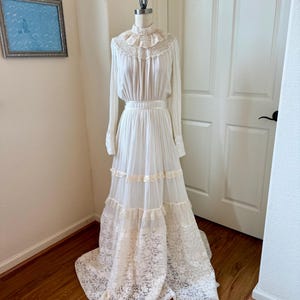 May include: A vintage white dress on a mannequin. The dress features a high ruffled collar, long sleeves, and a tiered skirt with lace and ribbon details. The dress is floor-length and appears to be made of a lightweight fabric.