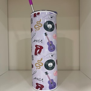 May include: A stainless steel tumbler with a purple lid and straw. The tumbler is covered in a pattern of musical and romantic symbols, including guitars, vinyl records, and the word "Lover". The background is a light purple color.