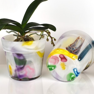 Puede incluir: Cuatro macetas decorativas de vidrio con base blanca y diseños abstractos coloridos en amarillo, rosa, verde y azul. Una maceta contiene una planta verde. Las macetas están dispuestas sobre una superficie blanca.