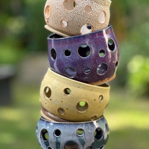 May include: A stack of four ceramic bowls in varying colors: beige, purple, yellow, and blue. Each bowl has a series of circular holes. The bowls are stacked on top of each other, with a blurred green background.