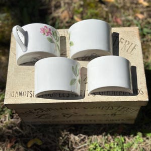 May include: Four white ceramic cups with floral designs, including pink flowers and green leaves. One cup has a handle. The cups are arranged on a wooden box with French text. The box is sitting on grass.