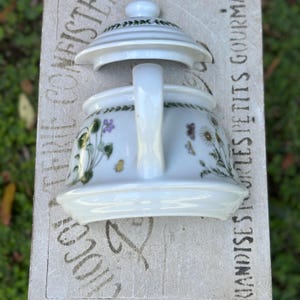 May include: White ceramic teapot with a floral design featuring purple and yellow flowers and green leaves. The lid is slightly ajar. The teapot is on a light-colored surface with the text "CONFISTERIE" and "FRIANDISES PETITS GOURMANDS".