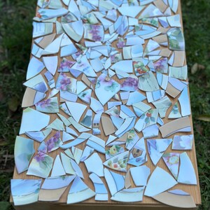 May include: A wooden box with a mosaic top made of broken china pieces. The china fragments feature floral patterns in shades of purple, pink, and green, set against a light blue and white background. The box is sitting on grass.