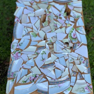 May include: A wooden table with a mosaic top made from broken china pieces. The china fragments feature floral patterns in pink, blue, and green, with gold trim. The table has a natural wood finish.