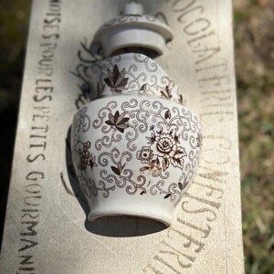 May include: A broken white ceramic pitcher with a brown floral design. The pitcher is sitting on a stone surface with the text "CANDISES POUR LES PETITS GOURMANDS" and "CHOCOLAT FIN ET DIGESTE" printed on it.