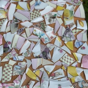 May include: A collection of broken porcelain pieces with floral, animal, and geometric patterns. The fragments feature pink, yellow, and white hues with gold trim. The pieces are arranged on a wooden surface, creating a mosaic effect.