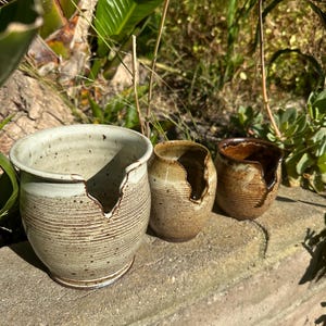 May include: Three ceramic pots with a crack in the side. The pots are in various shades of brown and white with a speckled glaze. The pots are sitting on a brick wall.
