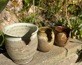 May include: Three ceramic pots with a crack in the side. The pots are in various shades of brown and white with a speckled glaze. The pots are sitting on a brick wall.