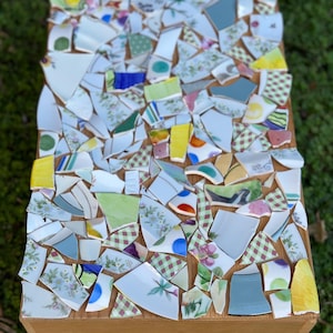 May include: A wooden box with a mosaic top made of broken pieces of colorful ceramic tiles. The tiles are arranged in a random pattern and include shades of blue, green, yellow, and white.