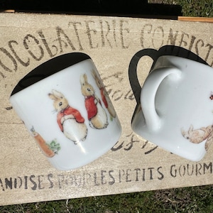 May include: Two white ceramic mugs with illustrations of Peter Rabbit and his friends. The mugs are on a wooden surface with the text "Chocolaterie Confiserie" and "Friandises pour les petits gourmands".