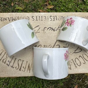 May include: Three white ceramic mugs with floral designs. The mugs have a curved shape and a handle on the side. The floral designs feature pink flowers and green leaves.