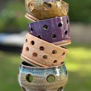 May include: A stack of four ceramic bowls in shades of brown, purple, and blue. The top bowl has heart-shaped cutouts. The other bowls have circular cutouts. The bowls have a textured, glazed finish.