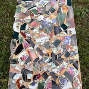 May include: A wooden table top mosaic made from broken pieces of decorative plates. The mosaic features a variety of colors and patterns, including floral designs, fruit motifs, and solid black and dark green sections. The table is rectangular and sits on a bed of green grass.