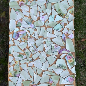 May include: A mosaic art piece made from broken china pieces. The fragments are arranged on a wooden board. The china has a light green background with floral designs in pink, yellow, and purple. The word "vera" is visible on one piece.
