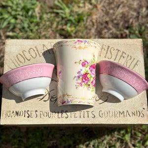 May include: Three vintage porcelain bowls with pink and white floral designs. The bowls are sitting on a wooden box with the text "Chocolaterie Fisterie Friandises pour les petits gourmands".