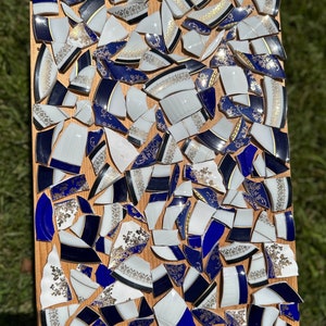 May include: A mosaic art piece made from broken pieces of blue and white china with gold accents. The pieces are arranged in a random pattern on a wooden background.