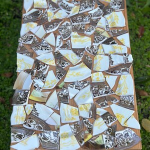 May include: A wooden table top mosaic made from broken china pieces. The china fragments are white, brown, and yellow, with floral patterns. The mosaic is set on a light brown wooden base, creating a unique decorative piece.