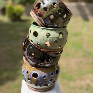 May include: A stack of four ceramic bowls with numerous circular holes. The bowls are in shades of green, brown, and blue, with a glossy finish. The bowls are stacked on top of each other, creating a unique and artistic display.