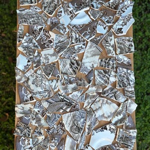 May include: A mosaic of broken ceramic pieces arranged on a wooden surface. The fragments feature black and white floral and landscape patterns. The overall design creates a unique decorative artwork.