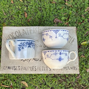 Puede incluir: Tres tazas de té de cerámica blanca con diseños florales azules y asas dispuestas sobre una superficie rectangular desgastada. Las tazas son de diferentes formas y tamaños. La superficie tiene texto descolorido: "Chocolaterie" y "Friandises pour les petits gourmands."