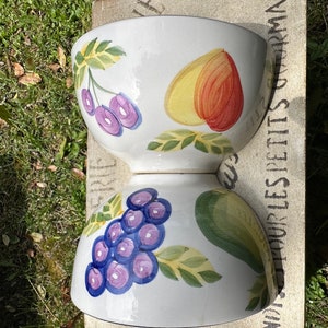 May include: Two white ceramic bowls with a fruit design. The top bowl has a peach, cherries, and green leaves. The bottom bowl has grapes, a green leaf, and yellow leaves.
