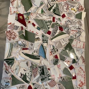 Puede incluir: Una obra de arte en mosaico hecha de piezas rotas de platos de cerámica. Las piezas están dispuestas en un patrón aleatorio y presentan varios colores, incluyendo blanco, verde, rojo y azul. La obra de arte está sobre un fondo blanco.