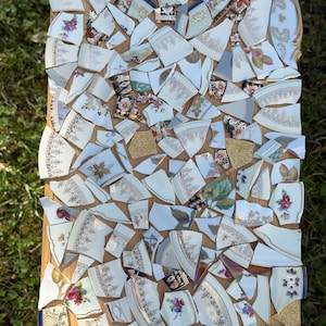 May include: A mosaic tabletop made from broken china pieces. The fragments are primarily white with floral and gold patterns, arranged on a wooden surface. The mosaic includes various shapes and sizes of china shards.