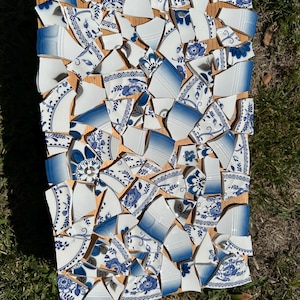 May include: A mosaic artwork composed of broken blue and white porcelain pieces arranged on a wooden board. The porcelain fragments feature floral and geometric patterns. The artwork is displayed outdoors on a bed of green grass.