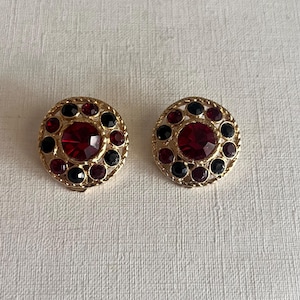 Op de afbeelding: Een paar goudkleurige clip-on oorbellen. Elke oorbel heeft een centrale, facetgeslepen, rode edelsteen omringd door kleinere rode en zwarte stenen. De oorbellen hebben een gestructureerd, cirkelvormig ontwerp.