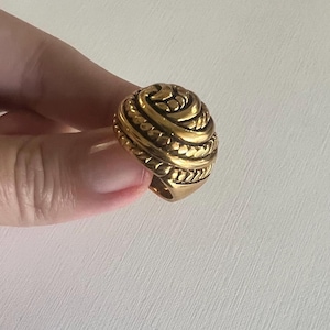 May include: A gold-toned ring with a textured, rope-like design. The ring is round and has a raised, intricate pattern.