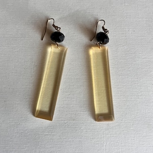 May include: A pair of dangle earrings featuring a black faceted bead and a long, rectangular, amber-colored pendant. The earrings have gold-colored ear wires and are displayed against a white background.