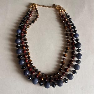 May include: A multi-strand necklace featuring three layers of beads in varying sizes and colors. The necklace includes dark blue, black, brown, and gold-toned beads. The necklace is fastened with a gold-colored clasp.