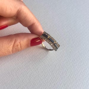 Pode incluir: Um anel retangular com uma banda dourada e um design de pedras pretas e transparentes. O anel é segurado entre os dedos com verniz de unhas vermelho. O design do anel apresenta uma pedra preta central ladeada por fileiras de pedras transparentes.