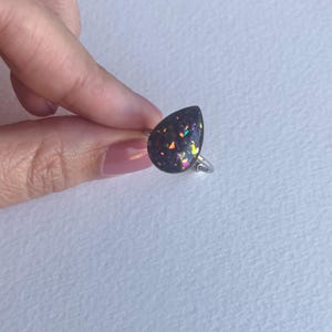 May include: A silver ring featuring a teardrop-shaped, dark-colored stone with iridescent flecks of color. The ring is held by a hand against a white background, showcasing the ring's design and sparkle.