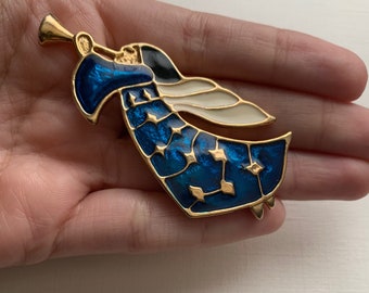 May include: A gold-toned angel brooch with blue enamel detailing and white wings. The angel is playing a trumpet. The brooch has a detailed design with gold accents and a classic, vintage style.