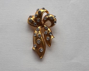 May include: A gold-toned brooch in the shape of a bow with clear rhinestones.