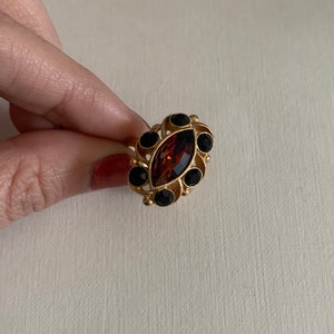 Pode incluir: Um anel de tom dourado com uma pedra central de cor âmbar em corte marquise, rodeada por seis pequenas pedras redondas pretas cravadas numa armação de metal decorativa com detalhes de volutas em torno da cravação central.