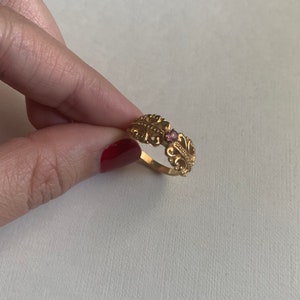 May include: A gold ring featuring an intricate filigree pattern along the band and a small, round, pink gemstone set in the centre. The metalwork displays detailed scrollwork and bead-like textures, creating a decorative band design.