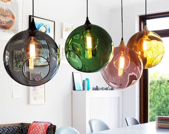 May include: Four spherical glass pendant lights hang from the ceiling, each featuring a different translucent colour: dark grey, green, pink, and amber. Each globe contains a single glowing Edison-style light bulb suspended by a black cord.