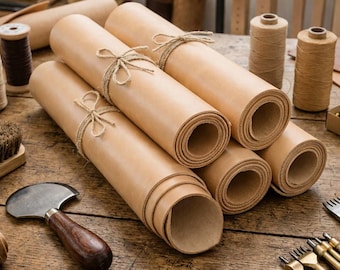 May include: Several rolls of light tan leather tied with thin jute twine. The leather rolls are arranged on a wooden surface alongside a metal head knife with a dark wooden handle and a set of metal craft punches and spools of thread.