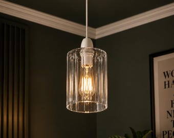May include: A cylindrical pendant light fitting featuring a clear, vertically ribbed glass shade suspended by a white cord and socket, housing a warm-toned Edison-style light bulb that emits a soft, glowing illumination.